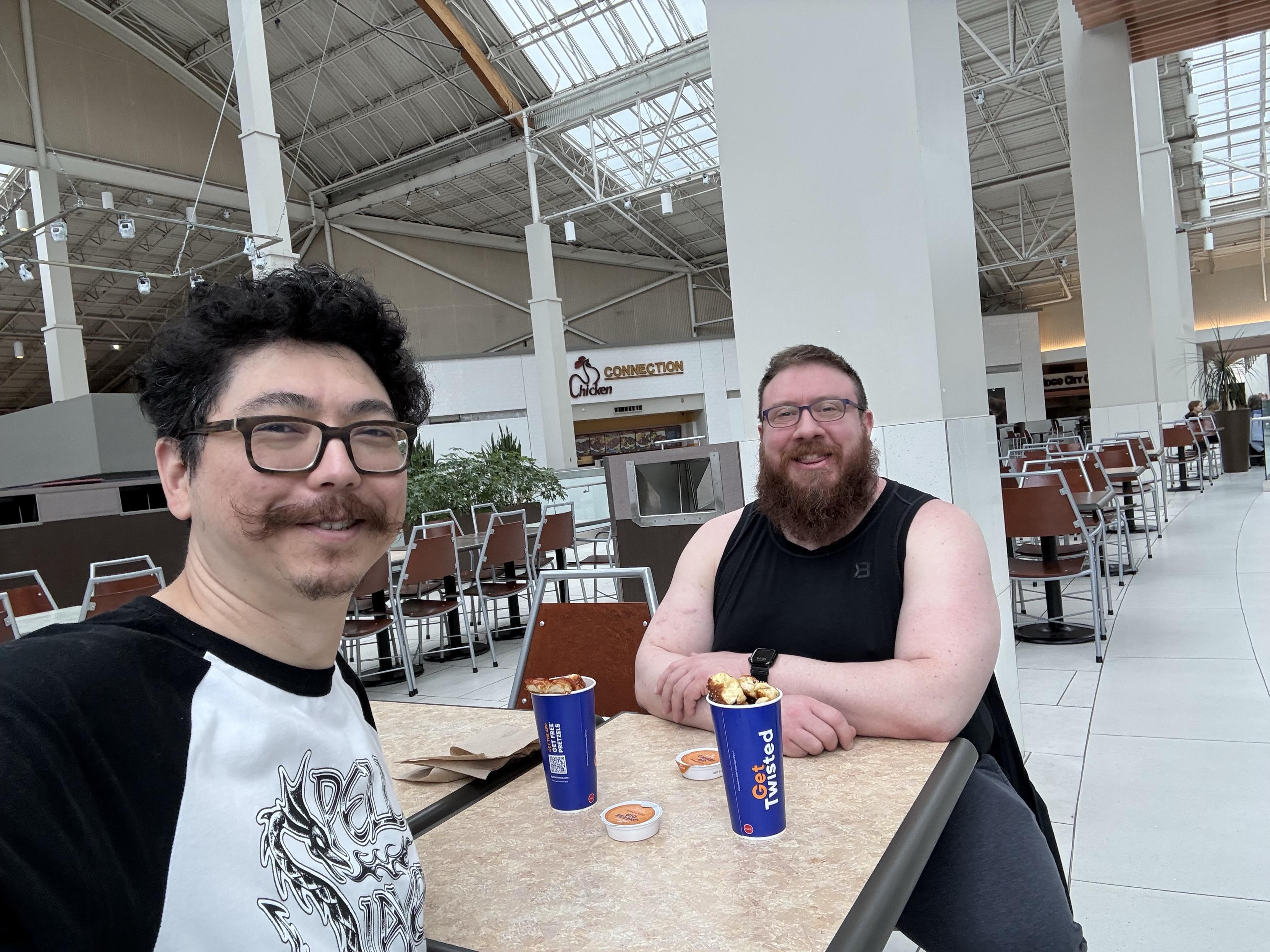 Two men are seated at a table in a food court area. Both are smiling; one has curly hair and a mustache, wearing a t-shirt, while the other has a beard and is wearing a sleeveless shirt. They have food containers in